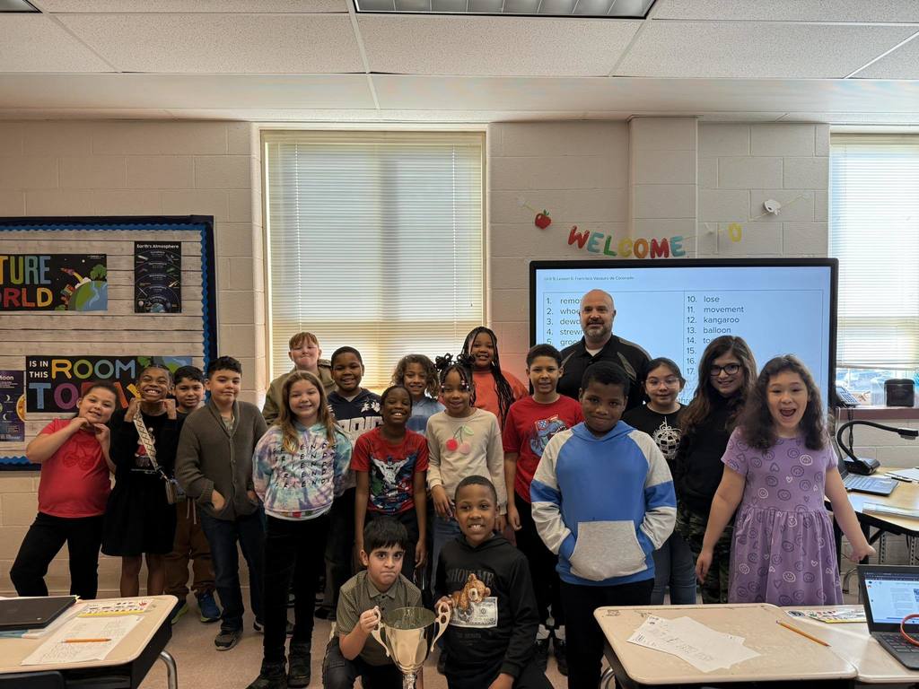Students and teacher posing in a classroom while one student holds a trophy. 