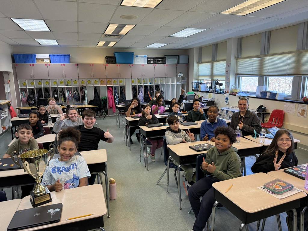 Students holding thumbs up at their tables in a classroom while a trophy is on one students' desk. 