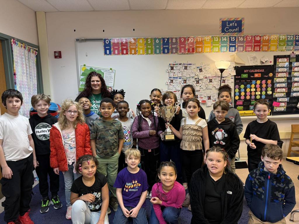 Students and teacher posing in a classroom while one student holds a trophy. 
