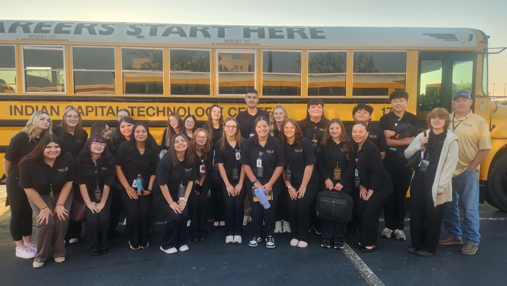 A group of students in matching uniforms, smiling, stands in front of a yellow school bus.