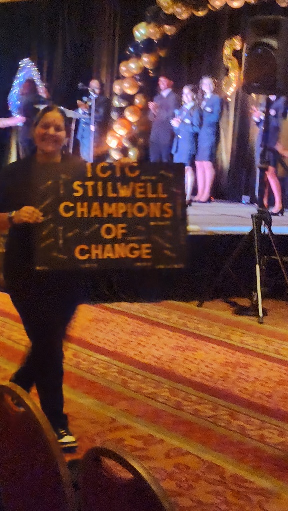 Person holds sign reading "ICTQ STILLWELL CHAMPIONS OF CHANGE" in front of a stage with people and balloons.