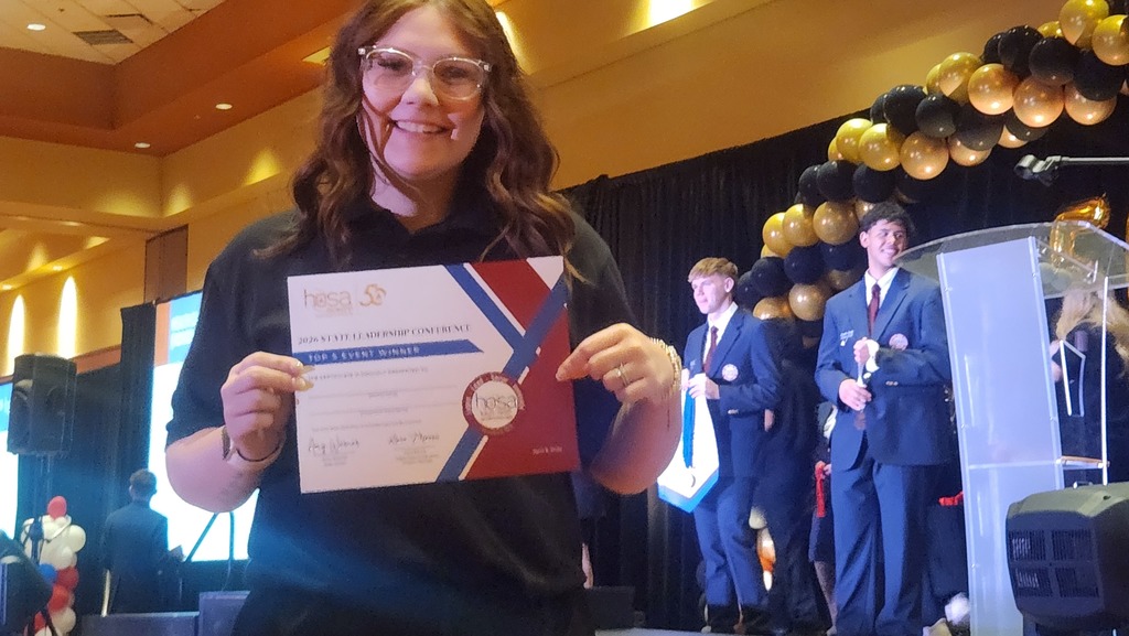 A woman in glasses holds a certificate with a medal at an event; three suited men stand behind her.