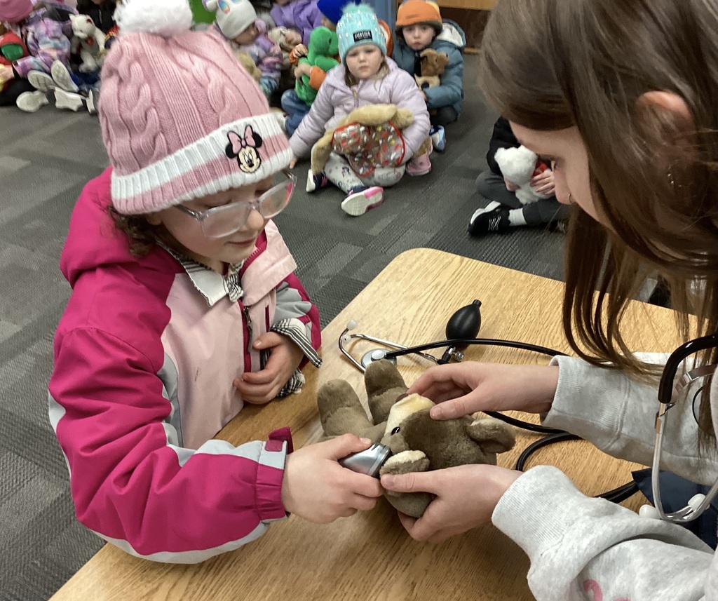 Student uses a toy medical tool on a stuffed animal at a classroom table while an adult assists; other students sit nearby with stuffed animals.