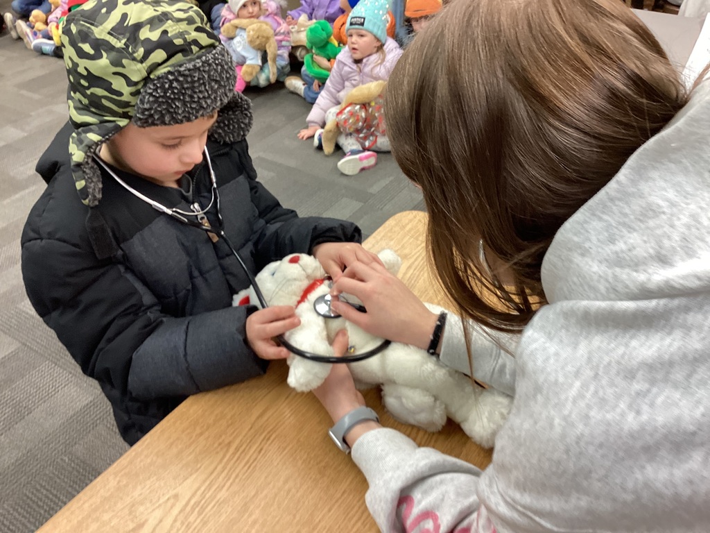 Student and adult use a stethoscope to “check” a white stuffed animal on a table during a teddy bear clinic activity.