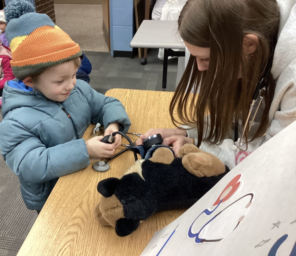 Student holds a stethoscope while an adult demonstrates checking a stuffed animal on the table.