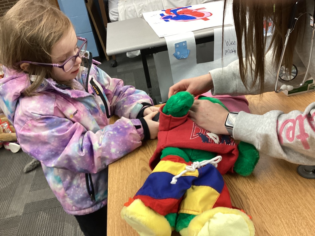  Student sits at a table while an adult helps examine a green stuffed animal dressed in bright clothing.