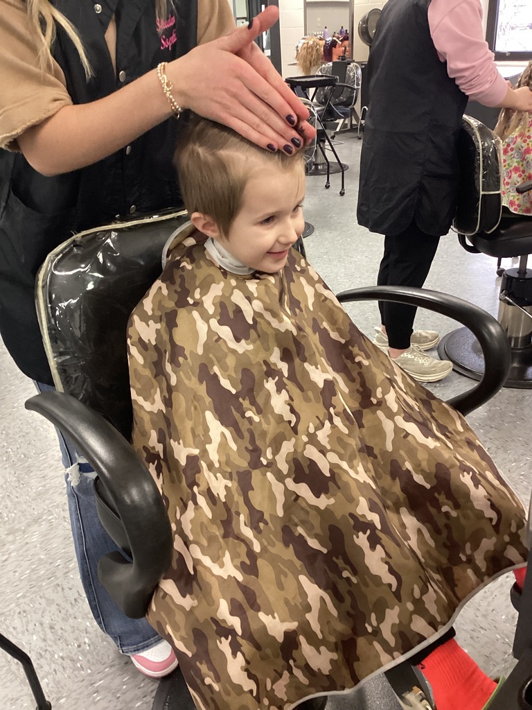 Cosmetology student smooths and styles a student’s hair while the student wears a camouflage haircut cape in a training salon.