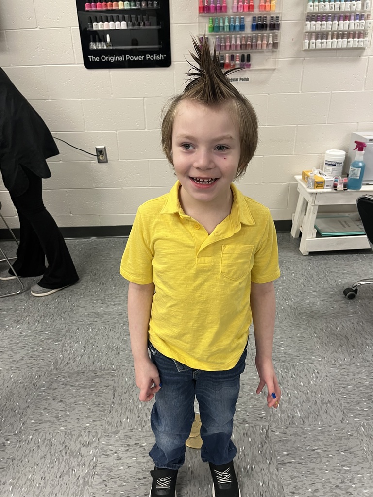  Student stands in the salon classroom with hair styled into a playful spiky look; nail polish displays hang on the wall behind.