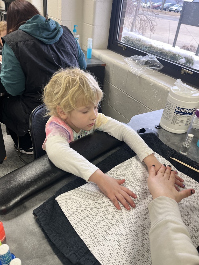  Student rests hands on a towel at a manicure station while an adult prepares to paint fingernails; nail supplies sit nearby.