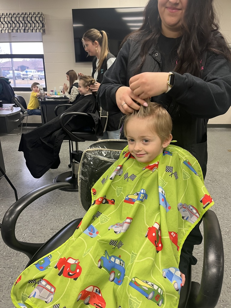 Cosmetology student styles a student’s hair while the student wears a bright green haircut cape; other salon stations appear in the background.