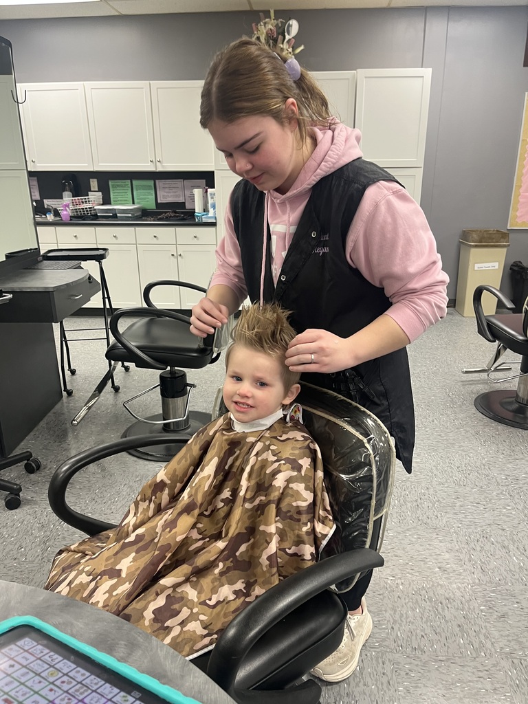 Student sits in a salon chair wearing a camouflage haircut cape while a cosmetology student styles their hair in a training salon.