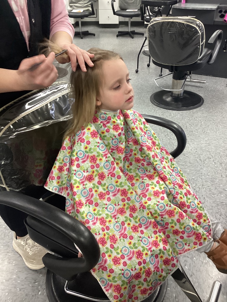 Cosmetology student brushes and styles a student’s hair while the student wears a floral haircut cape in a salon classroom.