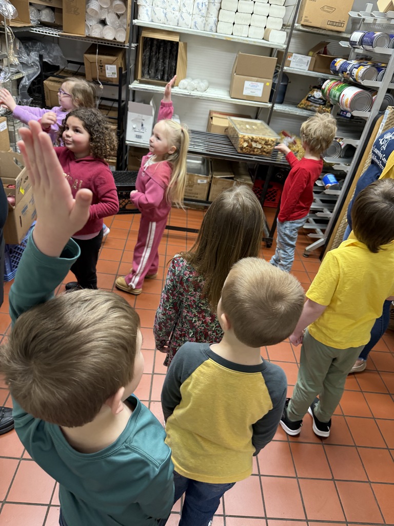  Students stand in a restaurant storage area with shelves of supplies while an adult talks and several students raise hands to participate.