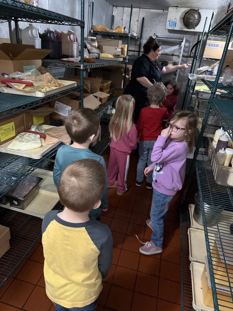 Students line up in a walk-in cooler while an adult points out items on shelves; metal racks and food containers are visible.