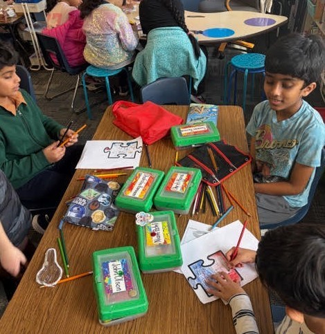 Kids seated around a table, engaged in a puzzle activity with pencils, papers, and a bag on the table.