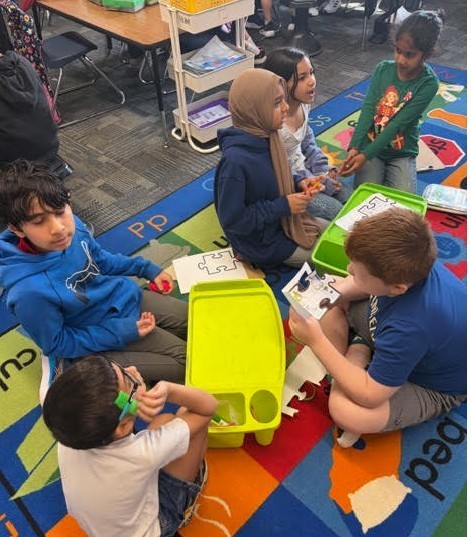 Children sitting on a rug, engaged in an activity with puzzles and materials, one child wears glasses.