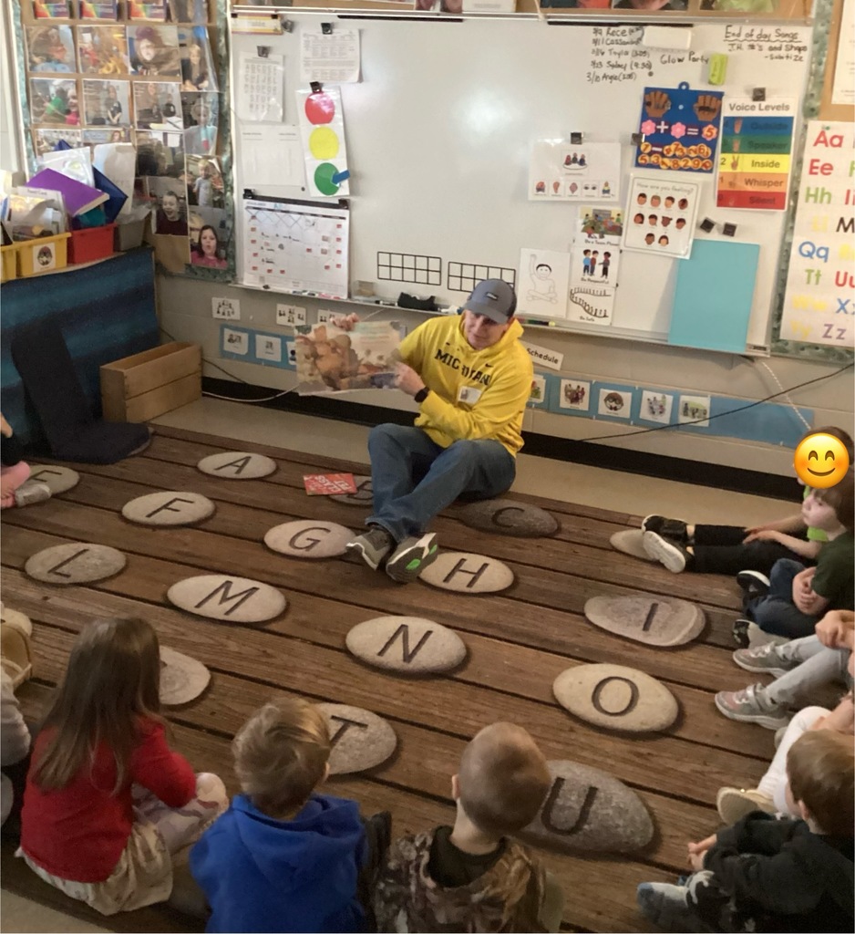 Adult guest reads a picture book aloud while students sit spaced around the classroom rug; classroom centers and learning materials are visible in the background.
