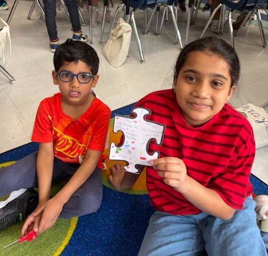 Two children sitting on a rug, holding a puzzle. One wears red striped shirt, the other in red shirt.
