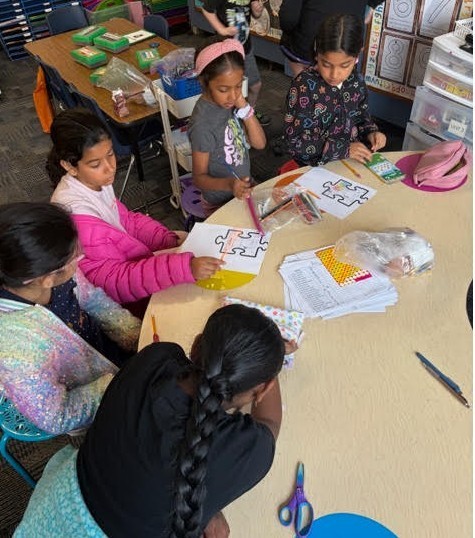 Five children, seated around a table, engage in a group activity involving paper puzzles, pens, and scissors.