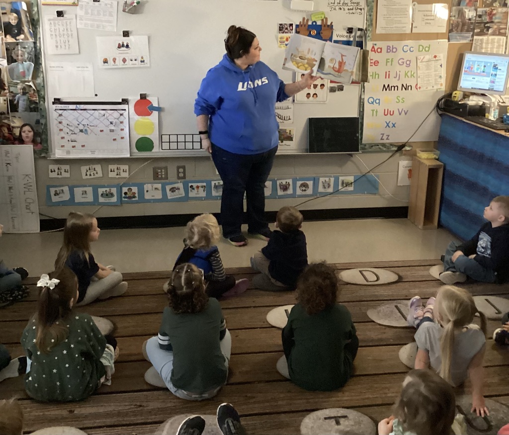 Adult guest stands at the front of the classroom holding an open book while students sit on the rug looking up during story time.