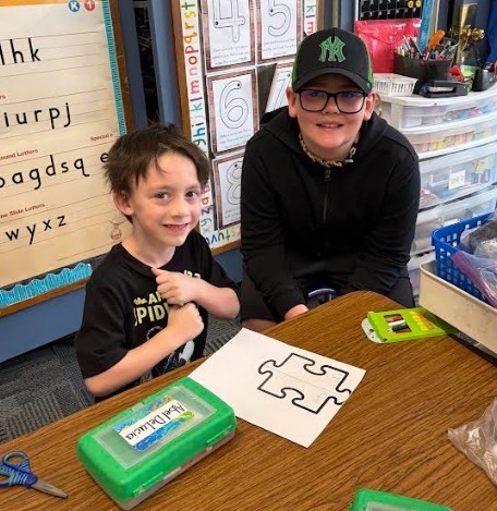 A boy smiles at the camera while seated at a desk. A man wearing glasses and a cap smiles beside him.