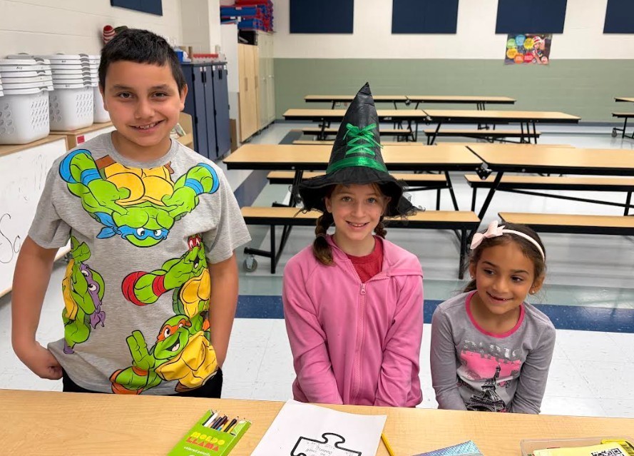 Three kids stand behind a table in a classroom, with one wearing a witch hat, another in a pink sweater, and the third in a gray shirt.