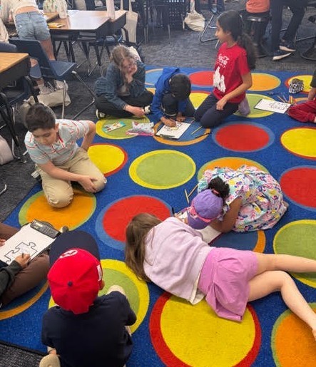 Kids sit in circle on a colorful rug, working on crafts. Some are seated on floor, others on chairs.
