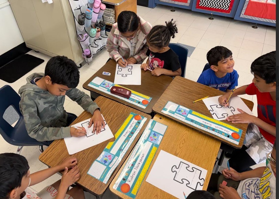 A classroom with children sitting at desks, working on puzzles and paper. Some have necklaces.