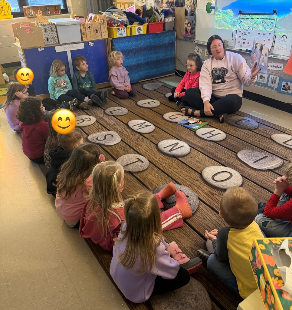 Adult guest sits on the classroom rug holding an open picture book while students sit in a circle listening; alphabet stones and classroom charts are visible.
