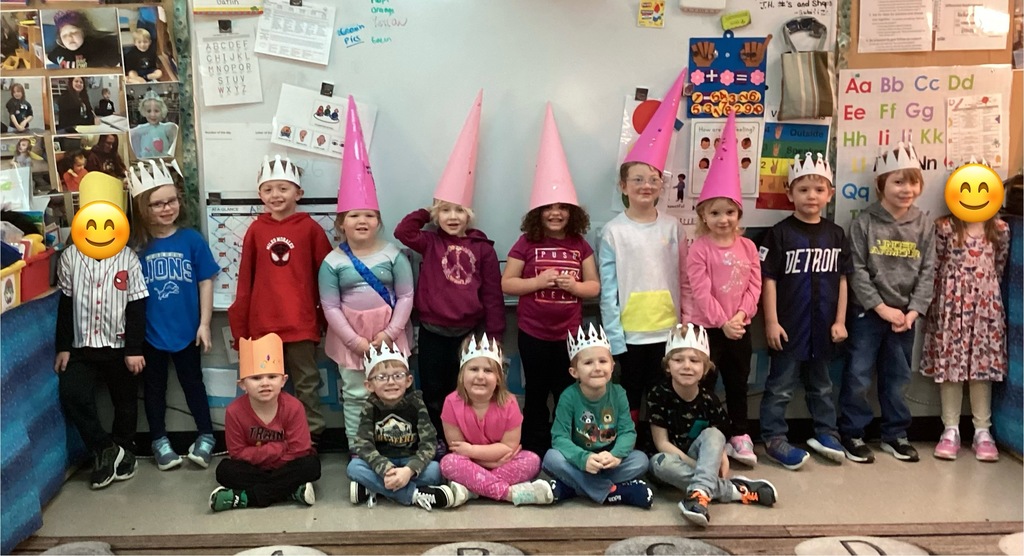 A large group of students pose together in the classroom wearing paper crowns and colorful cone hats during a themed classroom celebration; learning posters are visible behind them.