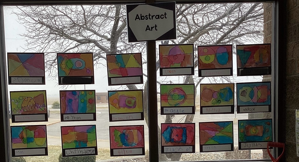 A classroom window display labeled “Abstract Art” shows multiple student artworks featuring colorful shapes, lines, and patterns arranged in neat rows.