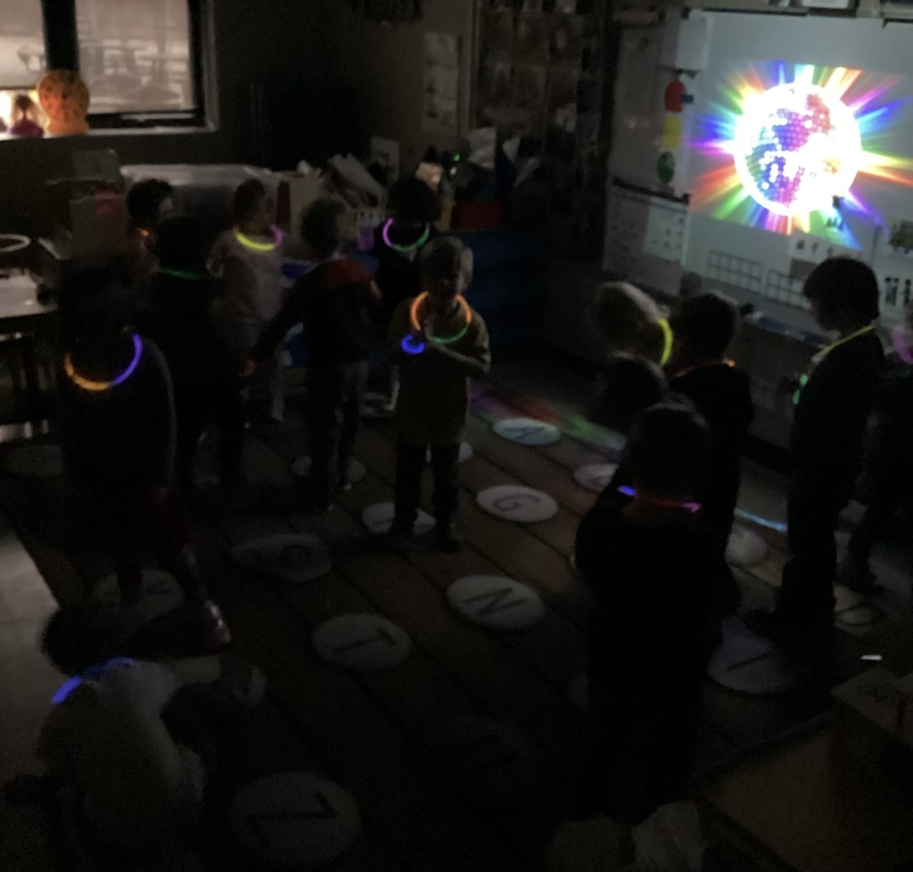 Students stand and move around the classroom during a glow party, wearing glowing necklaces while colorful light patterns shine on the wall in a darkened room