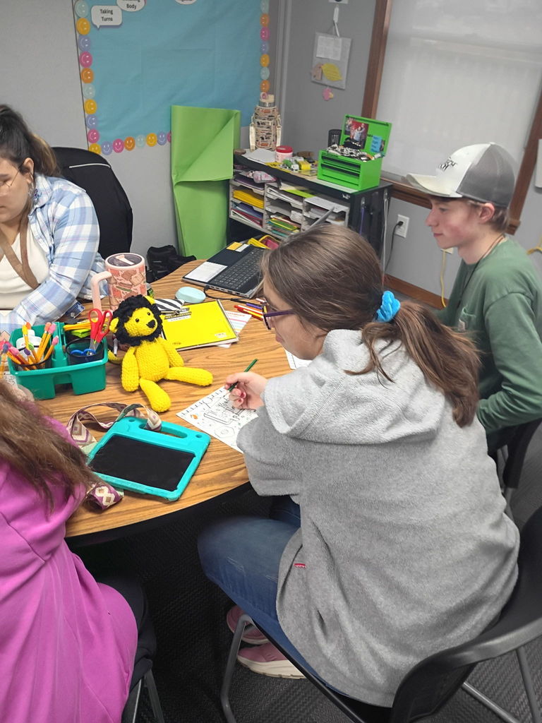 Secondary students sit around a table completing classroom activities with writing and art materials while a small crocheted lion sits in the center of the table as part of the lesson.