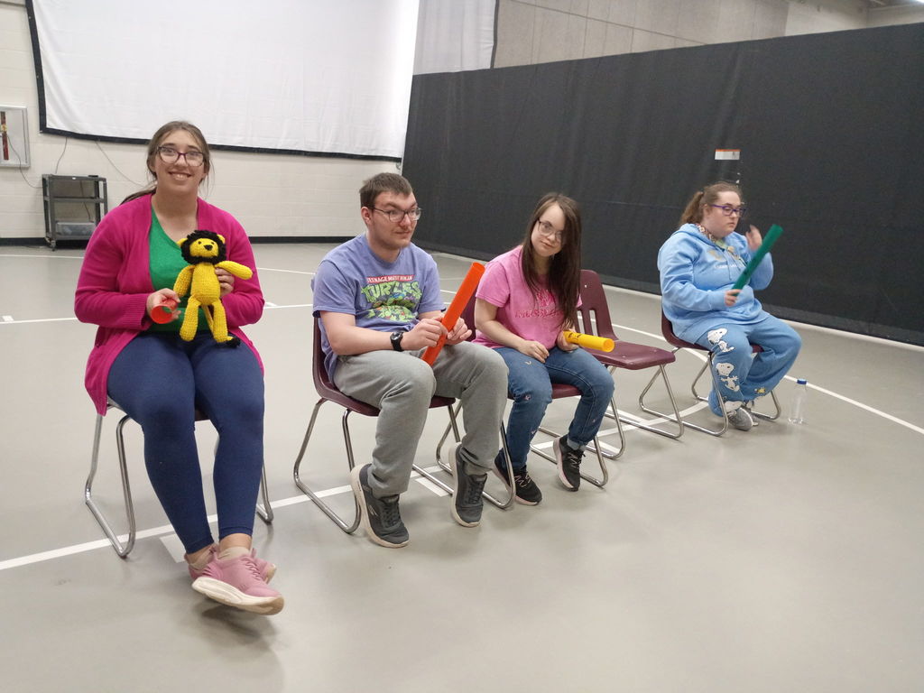 Secondary students sit in a row of chairs during a group activity while holding colorful rhythm tubes; a small crocheted lion is held by one student during the activity in a gym or multipurpose room.