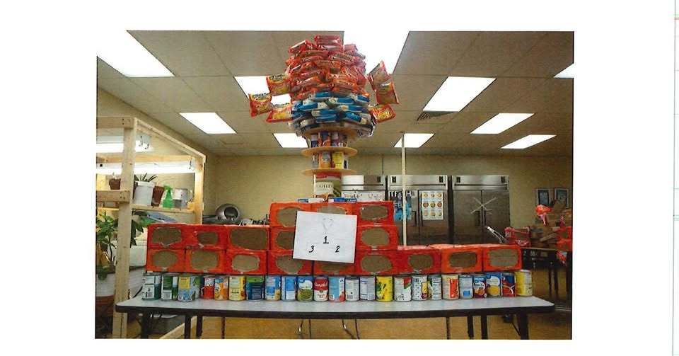 A large food sculpture displayed on tables inside a kitchen classroom, designed as a three‑tiered podium with a championship trophy on top. The structure is built from stacked boxed and canned food items, including pasta, soup, and packaged goods, arranged symmetrically. A paper labeled “1, 2, 3” is attached to the podium, and commercial kitchen appliances are visible in the background.