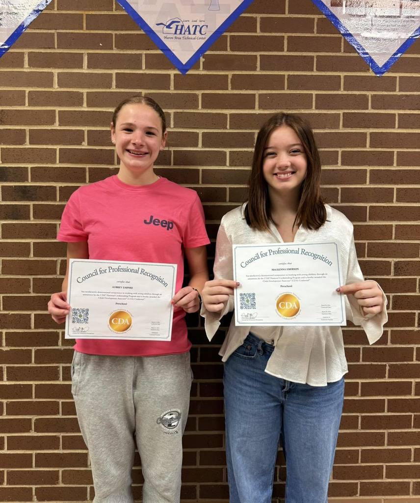 Two students stand in front of a brick wall holding certificates from the Council for Professional Recognition. Each certificate features a gold CDA seal, with a school banner displayed above them on the wall.