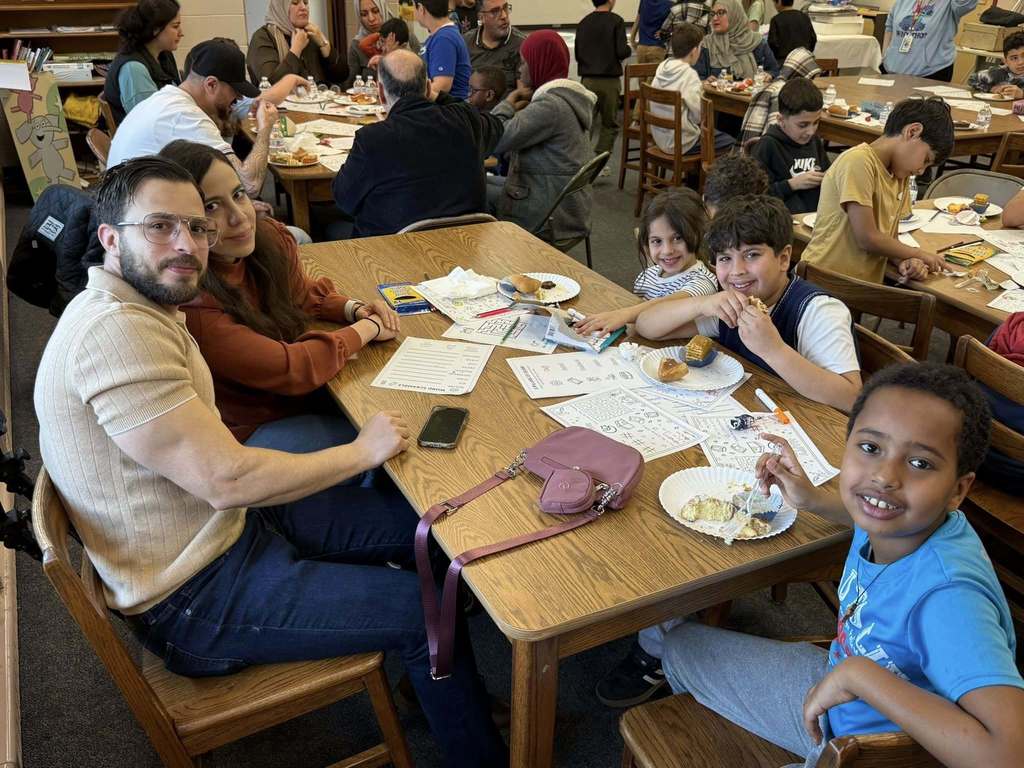 Students and two adults sitting at a table with paper and food in front of them. 