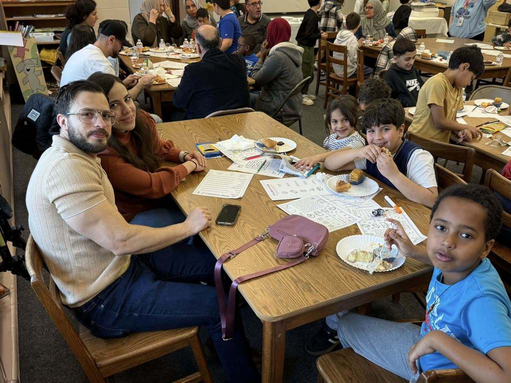 Students and two adults sitting at a table with paper and food in front of them. 