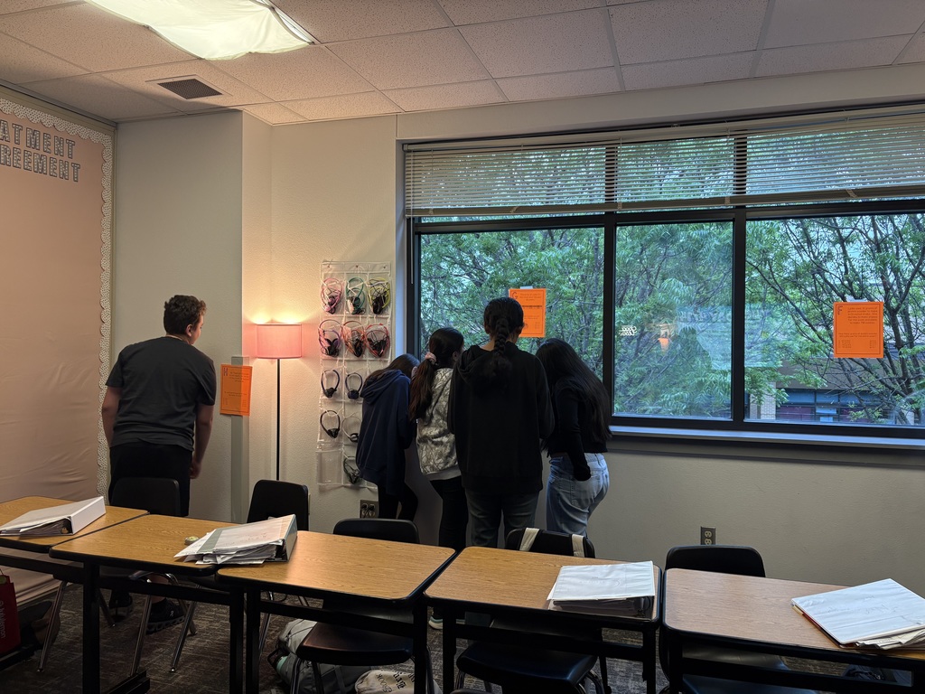 Several students move around the classroom during a STAAR review scavenger hunt, stopping at orange clue sheets posted on the wall and windows to solve problems about proportional relationships.