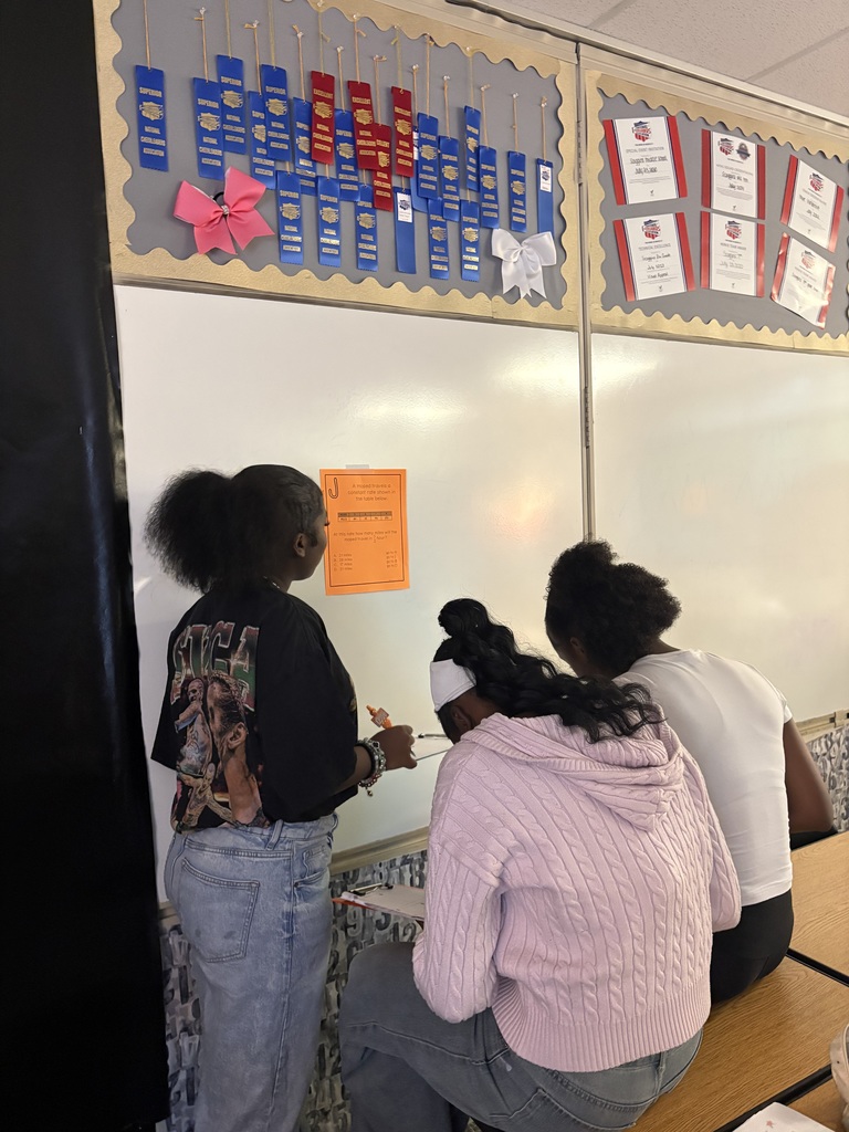 Three students work together at a classroom whiteboard during a STAAR review scavenger hunt on proportional relationships. One student stands and points toward an orange clue sheet taped to the board while the other two sit on a desk and write answers.