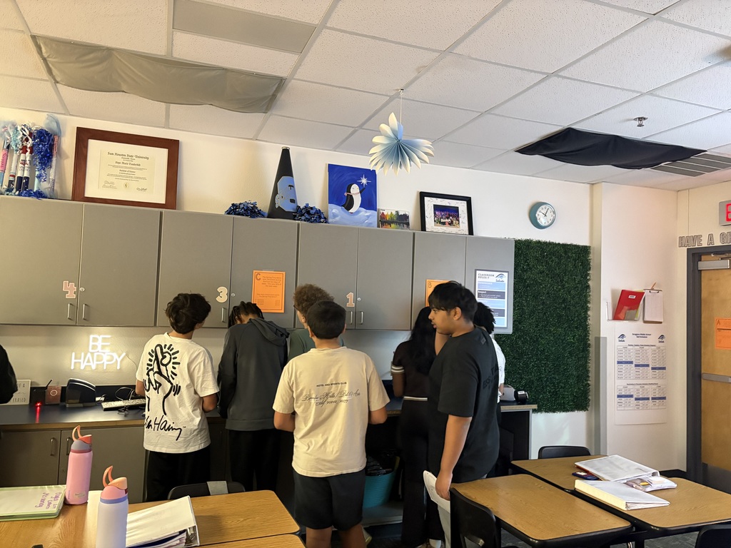 A group of students gathers around classroom cabinets during a STAAR review scavenger hunt, reading an orange clue sheet and working together to solve proportional relationship problems.