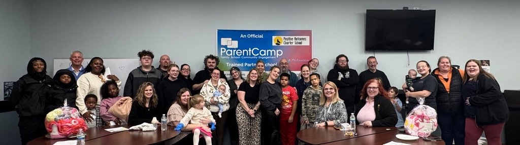 A group of people, adults and children, gathered indoors for a photo, standing behind tables, with a banner and TV in the background.