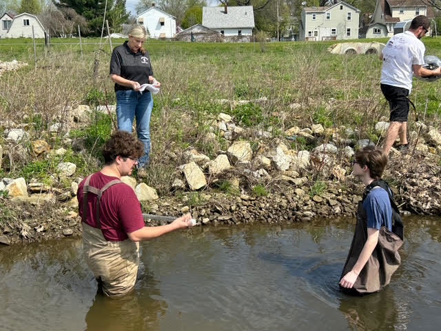 two students in the water with teacher Ms. Moran on the bank recording data