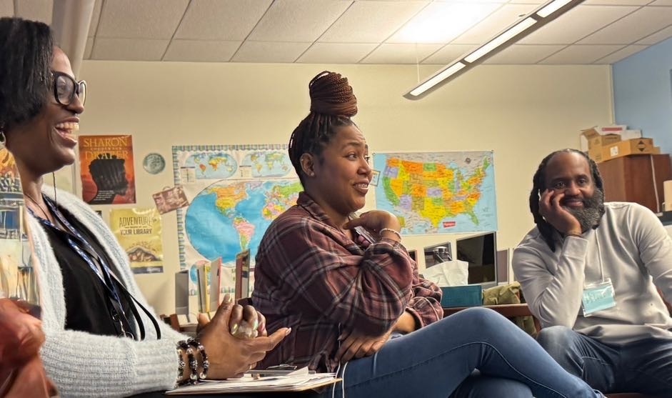 Three people sit together; two women and a man, smiling and speaking in a classroom.