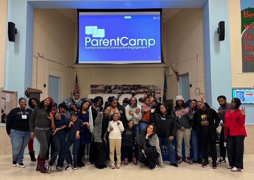 People gather in a room with a projector screen displaying the words "ParentCamp". Some wear winter clothing.