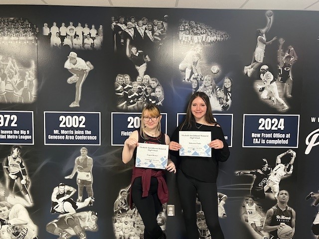 Two Students, Teagan Brewer (Left) and Makenzie Panks (Right) pose with thier student of the week certificates in front of a blue and white ahtletic themed mural.