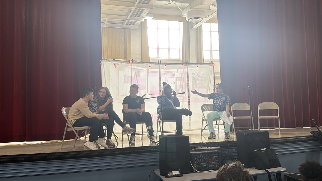 Students acting on stage while sitting. 