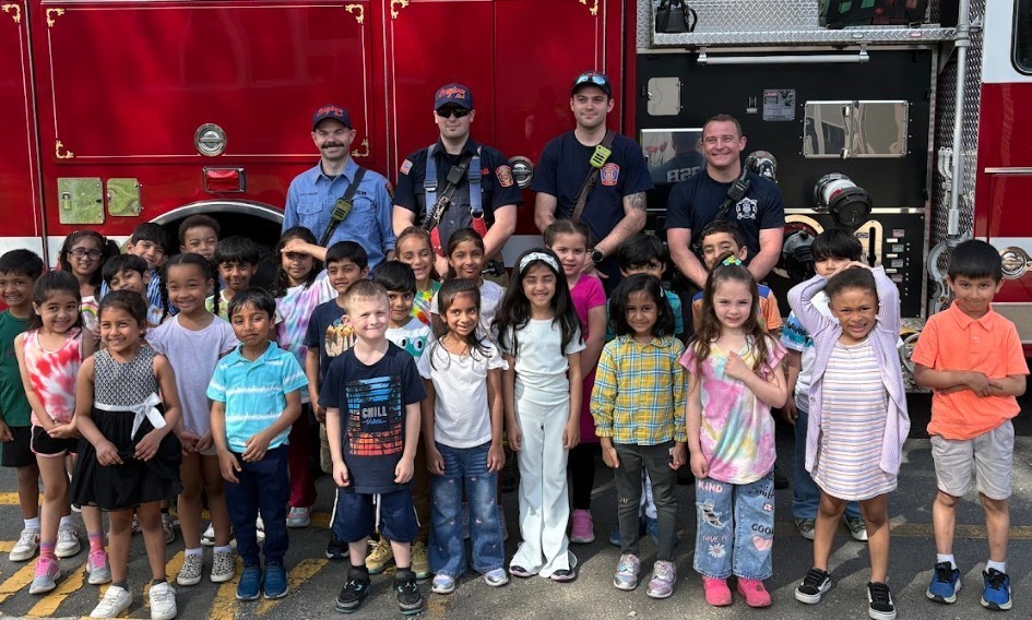 Group of children with firefighters in front of a red fire engine; children are dressed casually.