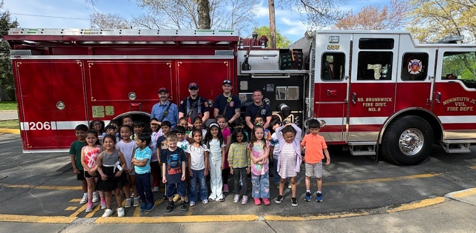 Group of children and two adult firefighters in front of a red fire truck in a parking lot.