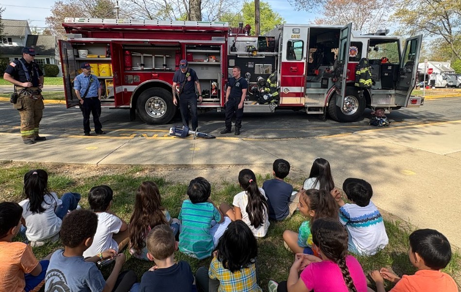 Kids sit on grass watching firefighters next to a red firetruck. One firefighter stands inside the truck.